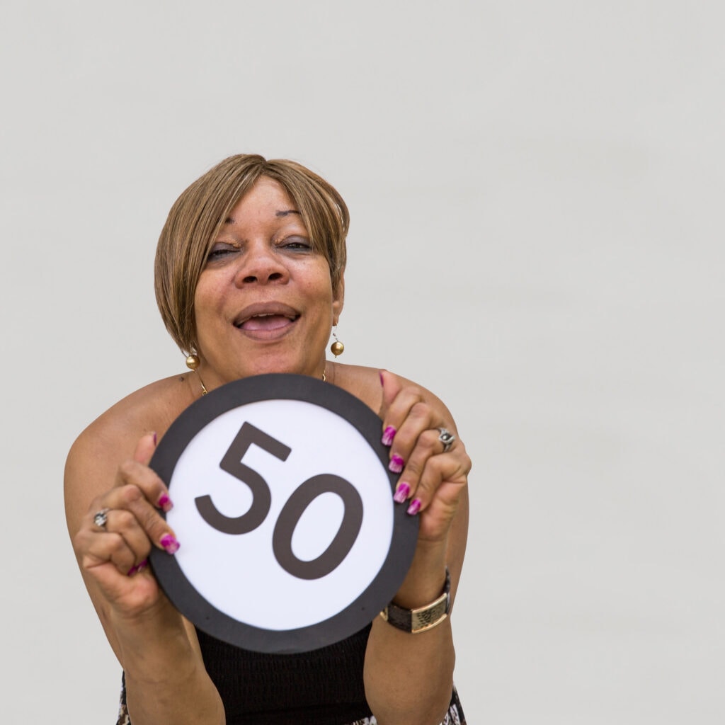 50 year old woman with an open smile laughs and looks into the lens with her eyes softly closing. With magenta painted fingernails she clutches a token in front of her body with the number 50 on it. This digital image is part of the 1 to Infinity portrait photography series by Danny Goldfield.