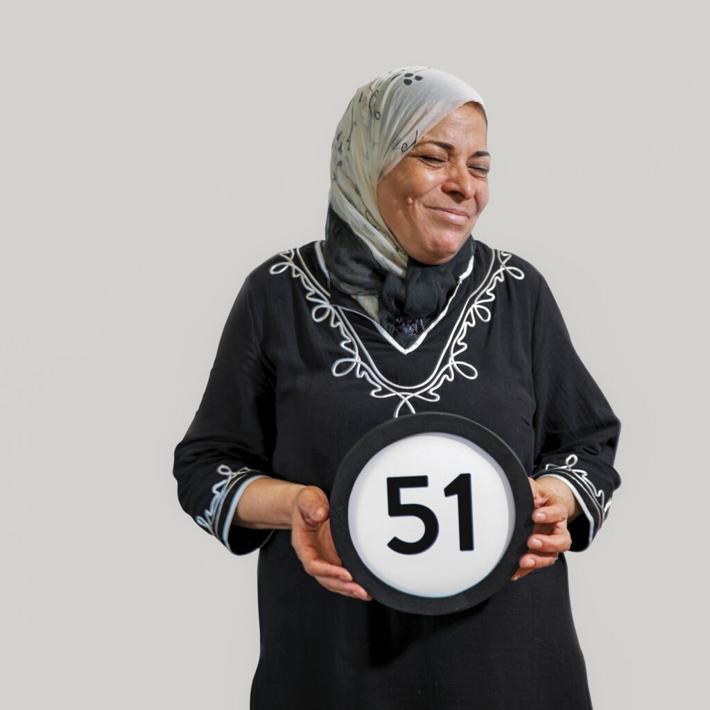 51 year old woman wears a scarf that covers her head and is tied around her neck. Her eyes are closed and she is laughing with her mouth closed. Her black dress has long sleeves and simple curvy embroidery design. She lightly holds a token with the number 51 on it. This digital image is part of the 1 to Infinity portrait photography series by Danny Goldfield.