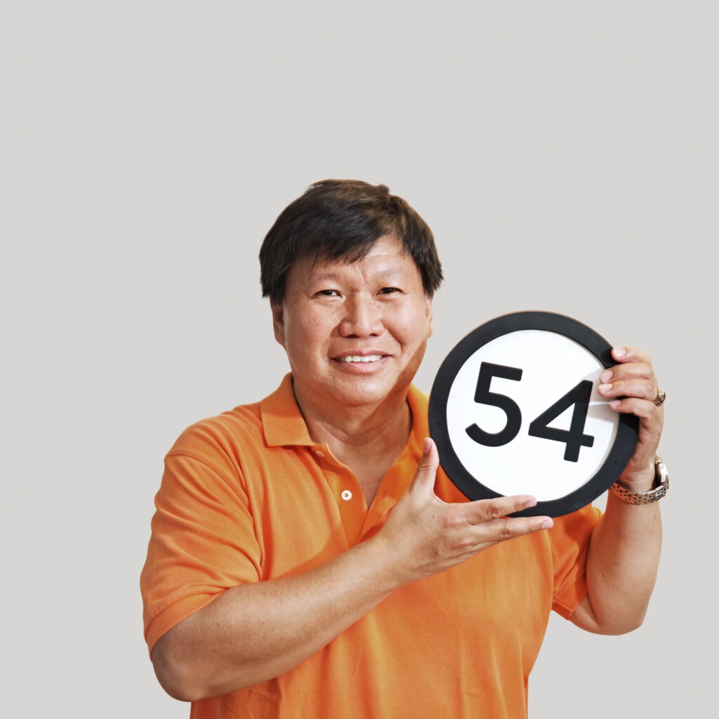 54 year old man wears a bright orange polo shirt and a relaxed smile that spreads across his whole face. His watch looks heavy and there is a college ring on his finger. In his hands just under his left cheek he holds up a token with the number fifty-four on it. This digital image is part of the 1 to Infinity portrait photography series by Danny Goldfield.