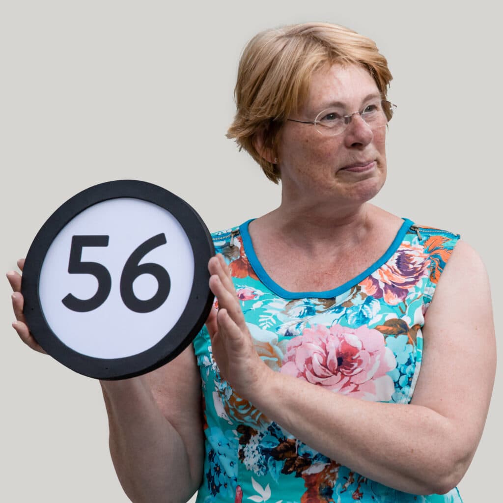 56 year old woman with short red hair and clear glasses wears a floral patterned dress with no sleeves. Her head is turned to her left with a terse but amused expression. To her right she holds up a token with the number 56 on it. This digital image is part of the 1 to Infinity portrait photography series by Danny Goldfield.