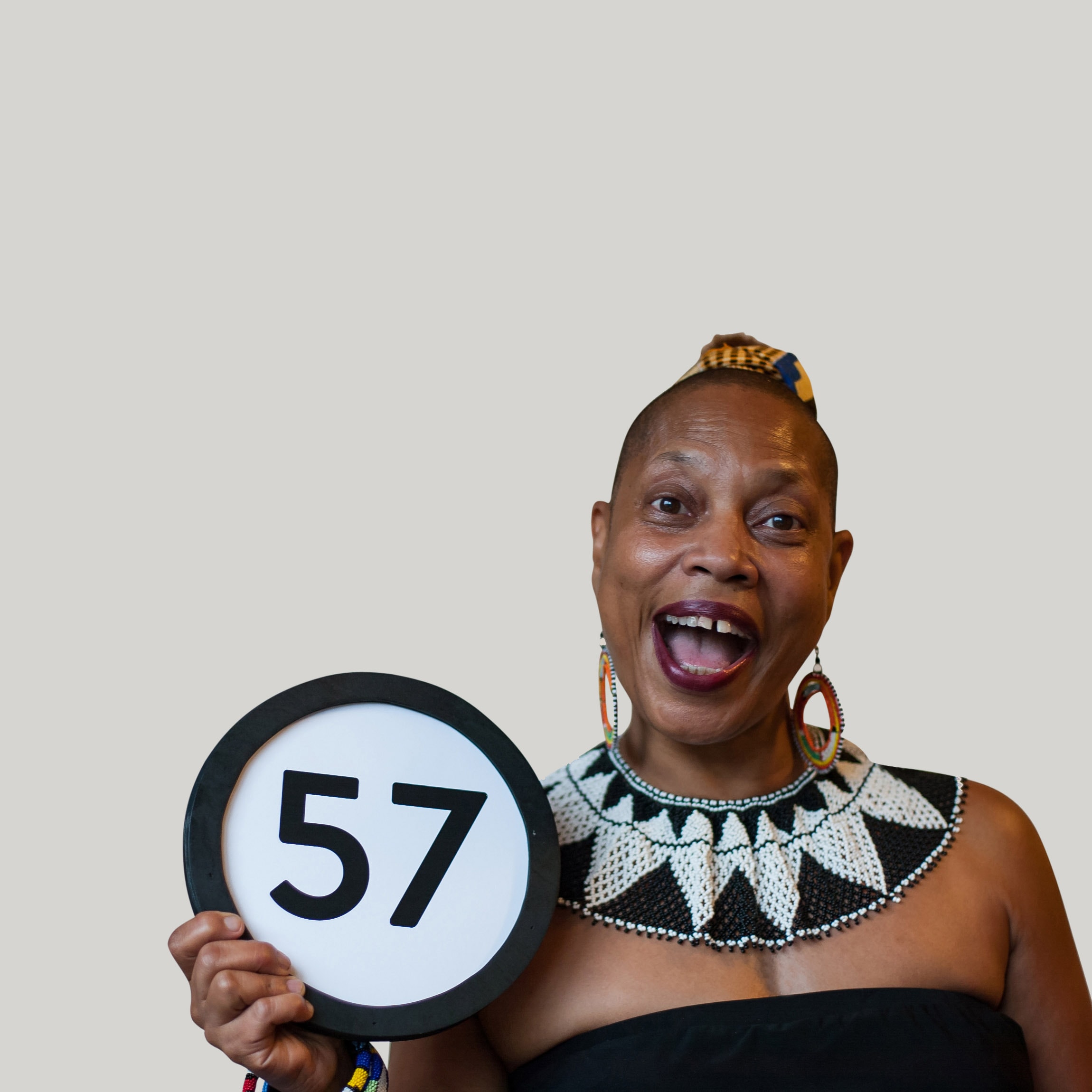 57 year old woman wears a black and white beaded collar, shoulderless dress, an open mouth smile. And she looks into the camera. Her head is shaved, lipstick is dark magenta, and she is thrilled to be holding a token with the number 57 on it. This digital image is part of the 1 to Infinity portrait photography series by Danny Goldfield.
