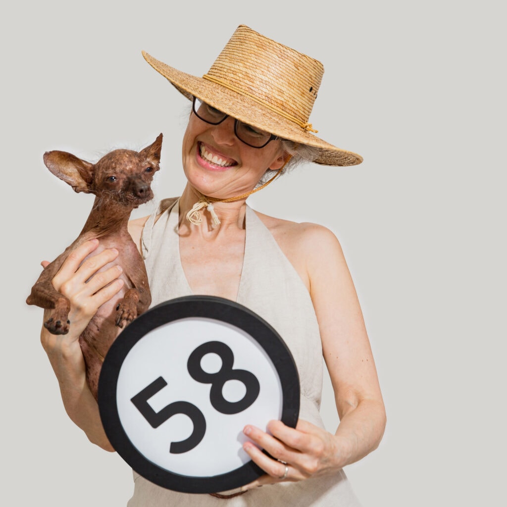 58 years old woman in a big straw sun hat smiles at her hairless alien dog that she holds in her right arm. The alien dog is looking into the lens. The woman also holds a token with the number 58 on it. This digital image is part of the 1 to Infinity portrait photography series by Danny Goldfield
