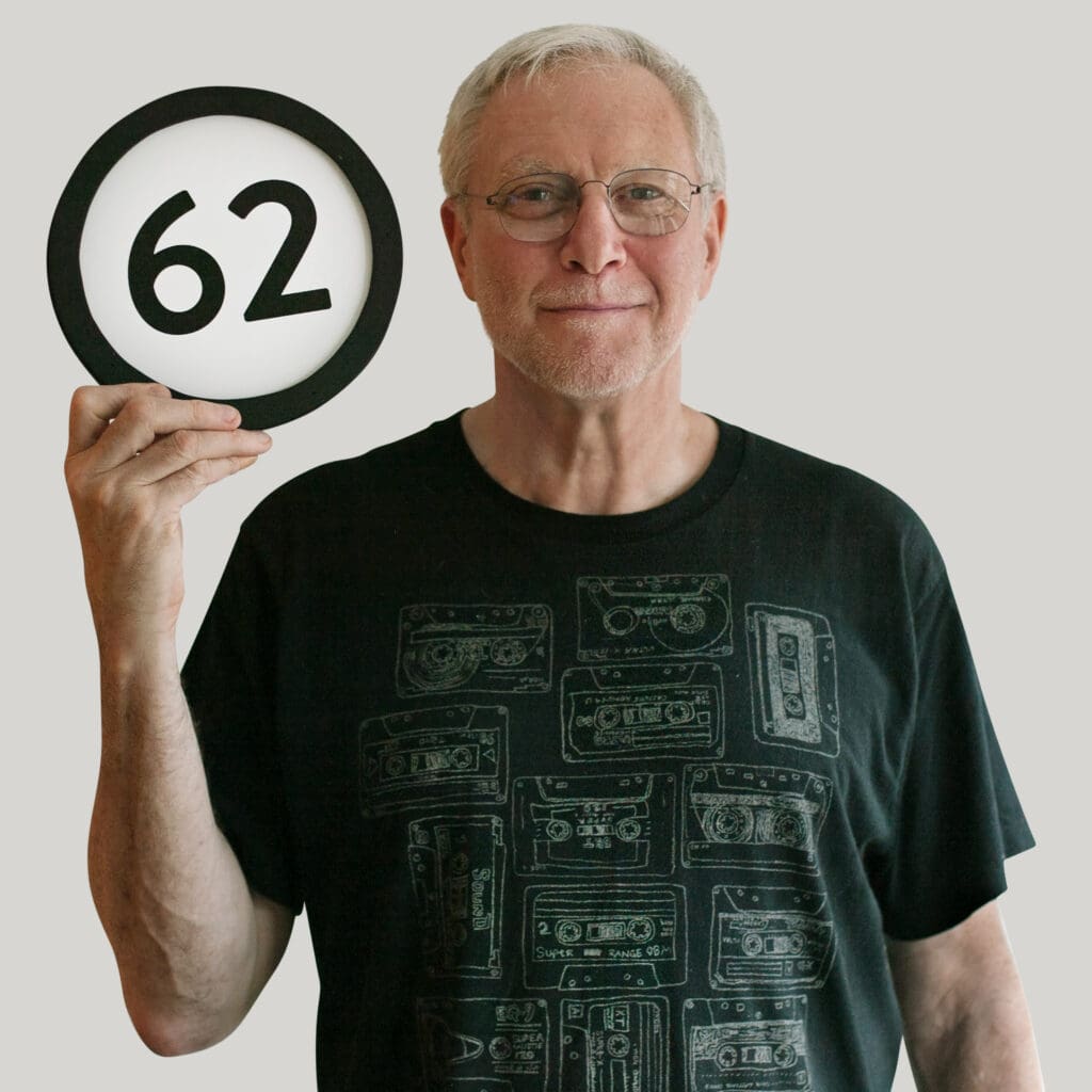 62 year old man wears a black t-shirt with a design of 13 sketches of music cassette tapes. He  has a calm closed smile and looks into the lens through his simple framed eyeglasses. In one hand he holds up a token with the number 62 on it. This digital image is part of the 1 to Infinity portrait photography series by Danny Goldfield