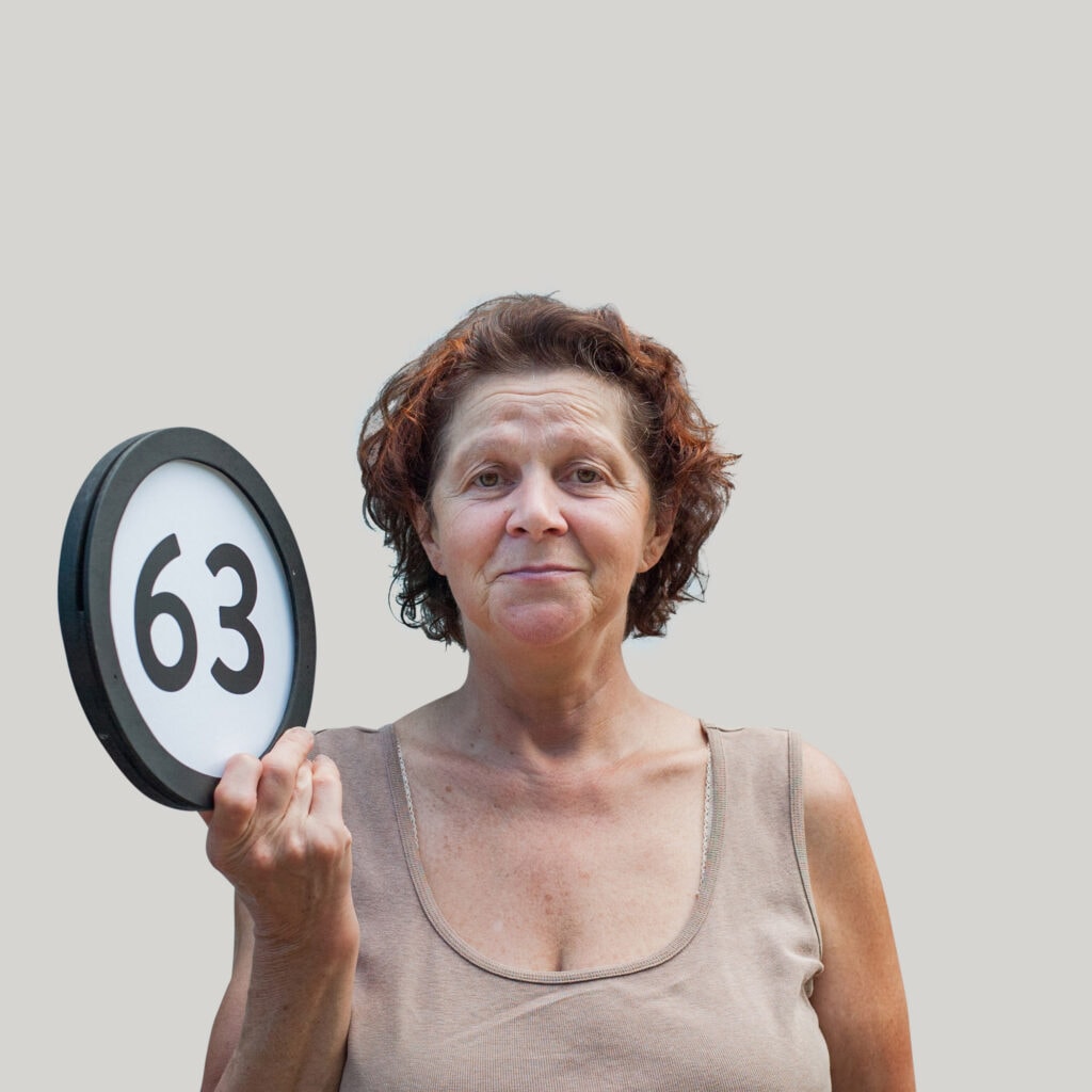 63 year old bemused woman holds with one hand a token with the number sixty-three on it. This digital image is part of the 1 to Infinity portrait photography series by Danny Goldfield..