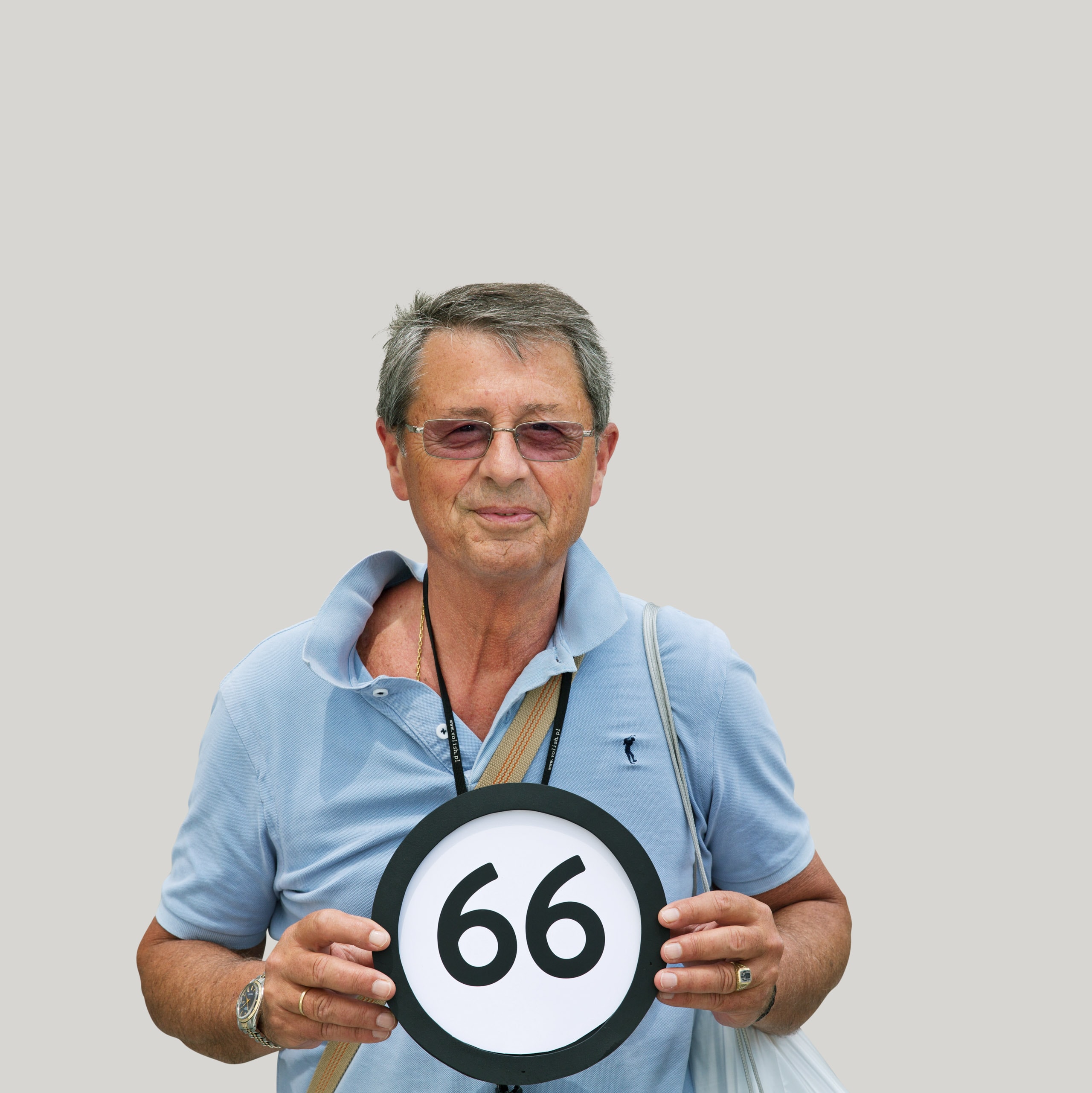 66 years old, tan, happy man in purple tinted sunglasses wears a light blue polo shirt with his collar unbuttoned and open. Straps of cotton tote bags are over each shoulder, he wears a fancy silver watch and two silver rings. With his fingertips he holds a token that has the number 66 on it. This digital image is part of the 1 to Infinity portrait photography series by Danny Goldfield.