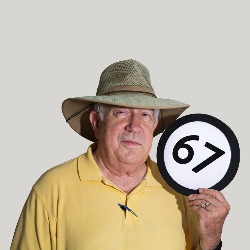 67 year old man wears a worn-in sunhat and faded yellow polo shirt. He looks into the lens with a relaxed expression. A silver Claddagh ring is on the hand that holds up a token with the number 67 on it. This digital image is part of the 1 to Infinity portrait photography series by Danny Goldfield.