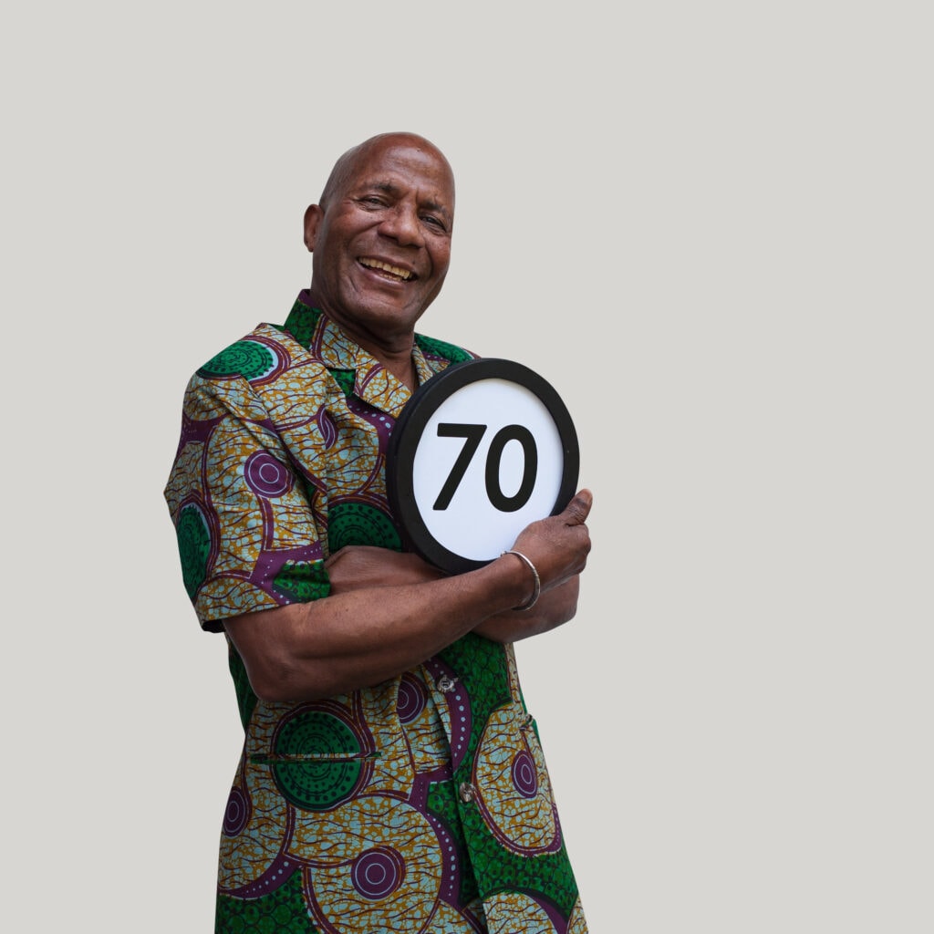 70 your old man wears a colorful summer shirt and smiles broadly into the lens. His arms are folded tightly to his chest and with one hand holds a token with the number 70 on it. This digital image is part of the 1 to Infinity portrait photography series by Danny Goldfield.