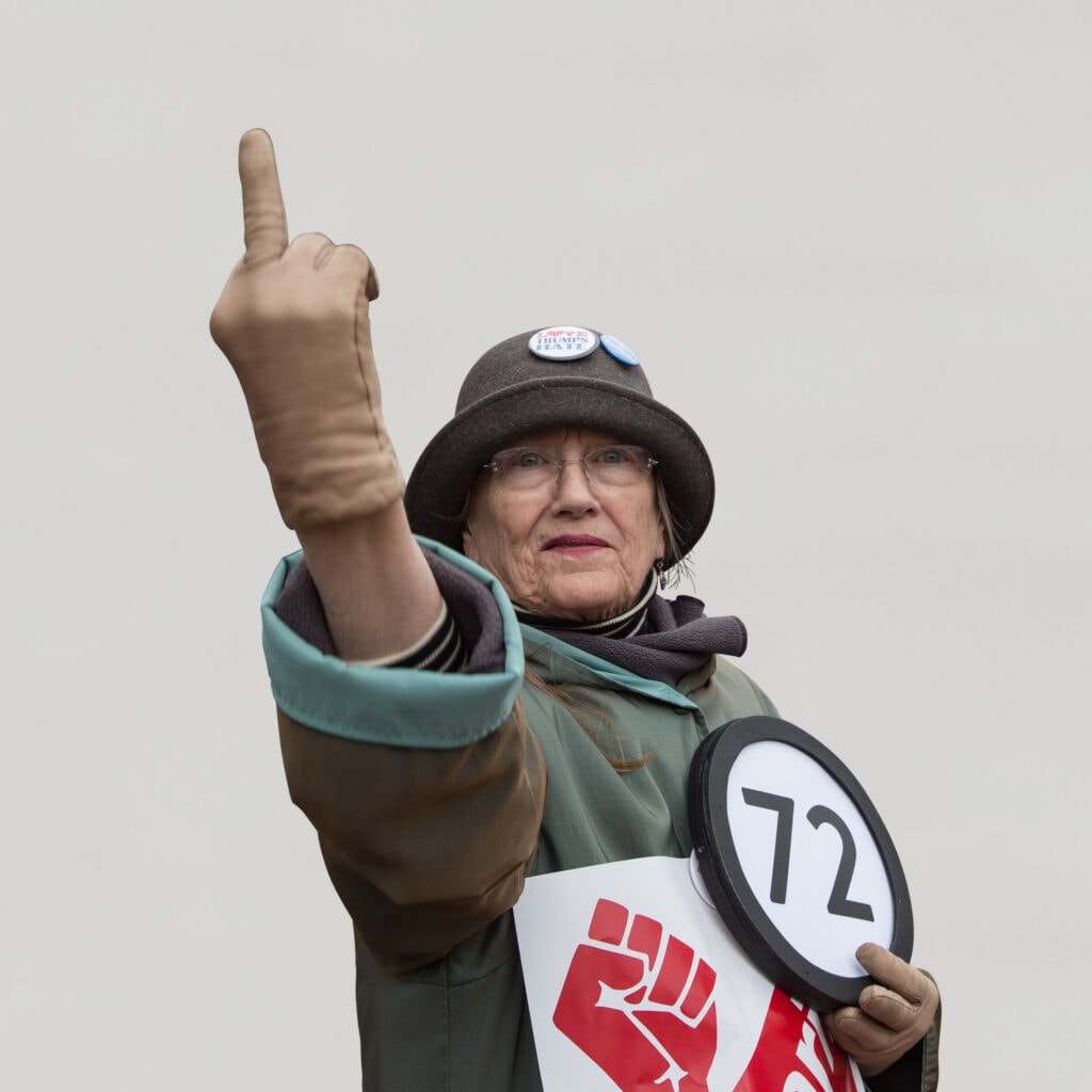 72 year old woman wears a "Love Trumps Hate" button on her wool cap and directs her middle finger towards the 45th president of the United States of America. Held  over her heart with her other hand is a token with the number 72 on it. This digital image is part of the 1 to Infinity portrait photography series by Danny Goldfield.