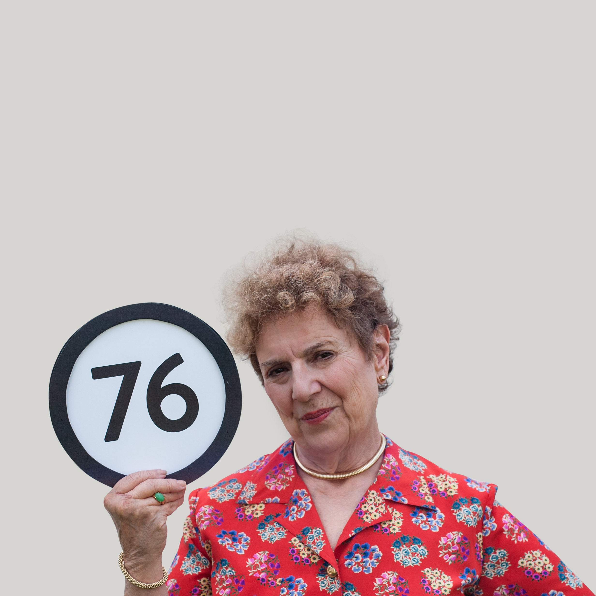 76 year old woman wears a red floral shirt, sleek gold jewelry, and a serious gaze into the lens. With one hand she grips a token with the number 76 on it. This digital image is part of the 1 to Infinity portrait photography series by Danny Goldfield.