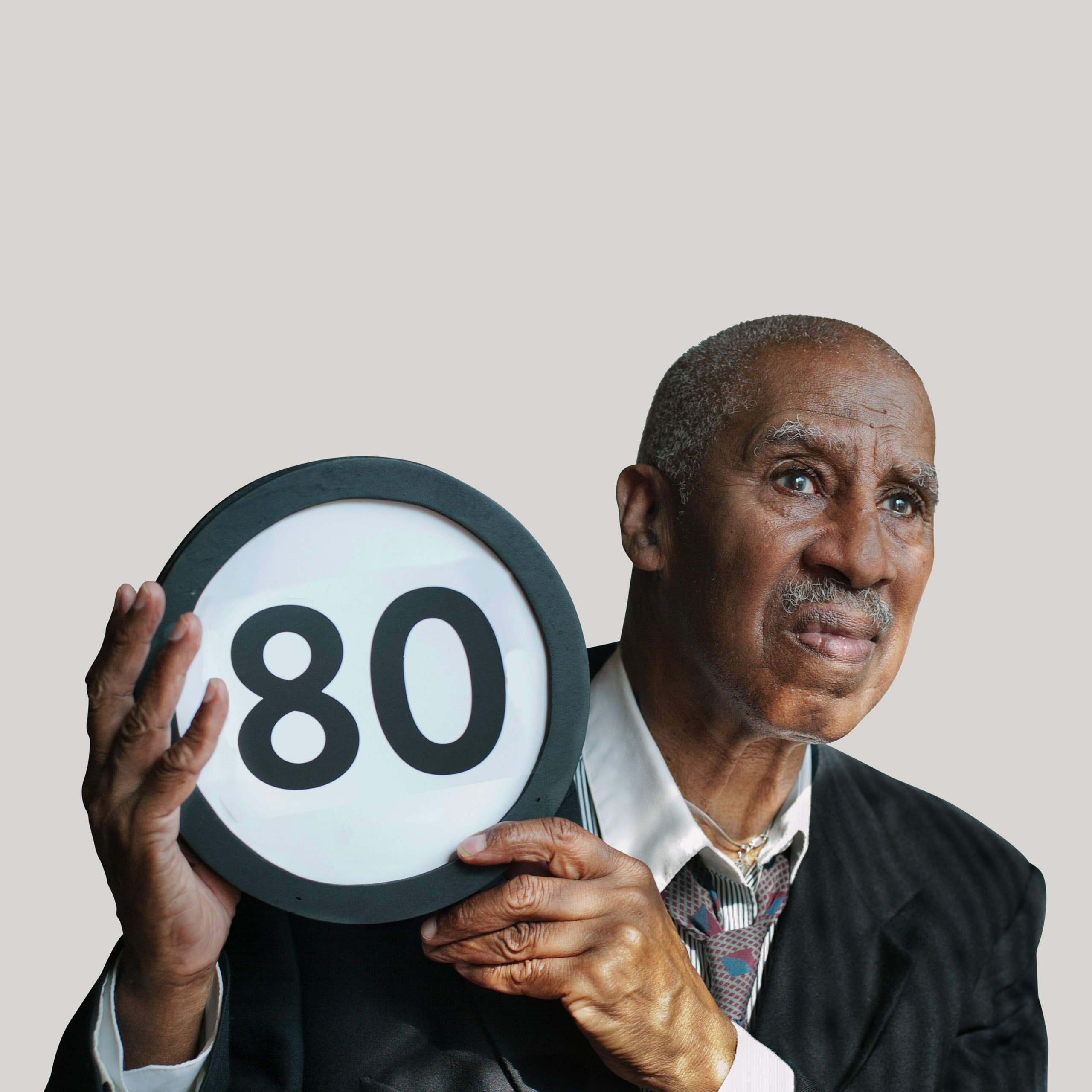 80 year old in an oversized suit jacket and loosened necktie looks concerned. He looks off into the distance while holding up a token with the number 80 on it. This digital image is part of the 1 to Infinity portrait photography series by Danny Goldfield.
