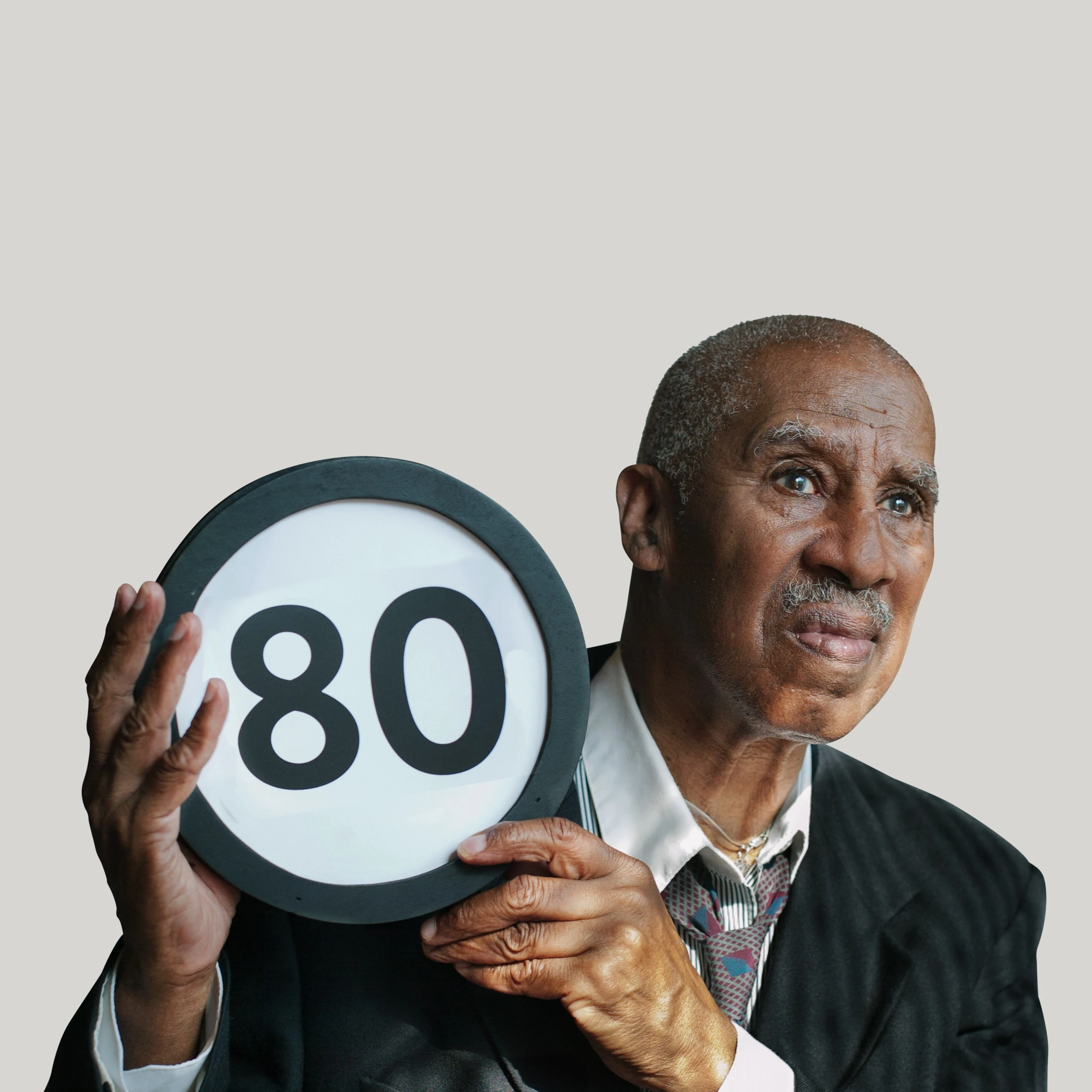 80 year old in an oversized suit jacket and loosened necktie looks concerned. He looks off into the distance while holding up a token with the number 80 on it. This digital image is part of the 1 to Infinity portrait photography series by Danny Goldfield.