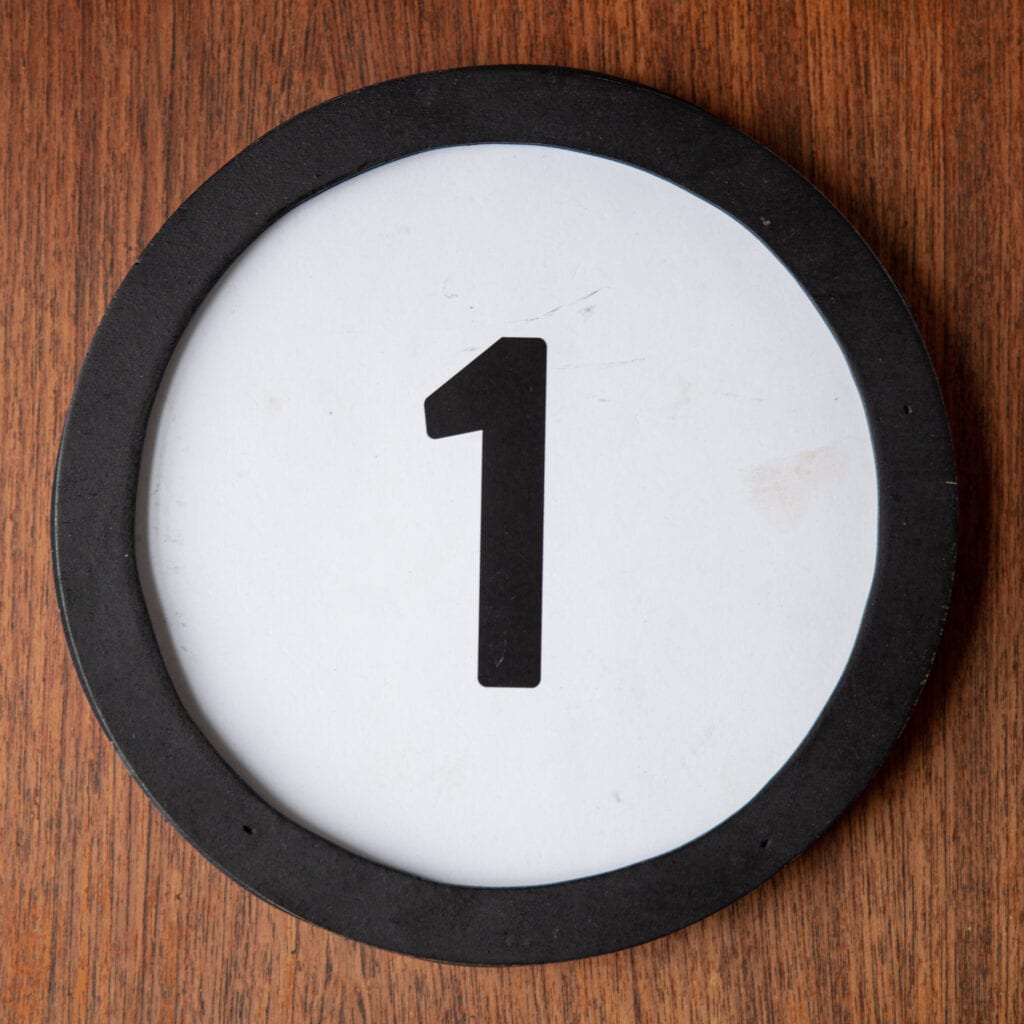 A circle shaped token fills a square image. The token has a 1 inch black circular frame around it. In the center is a black number on a white background. The token lies on a teak wood table. This digital image is part of the 1 to Infinity portrait photography series by Danny Goldfield.