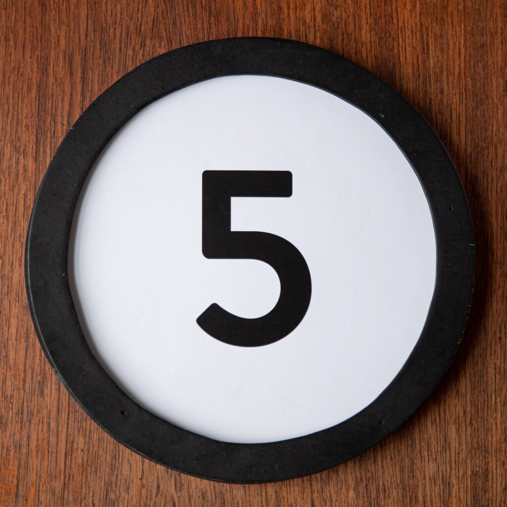 A circle shaped token fills the square image. The token has a 1 inch black circular frame around it. In the center is a black number on a white background. The token lies on a teak wood table. This digital image is part of the 1 to Infinity portrait photography series by Danny Goldfield.