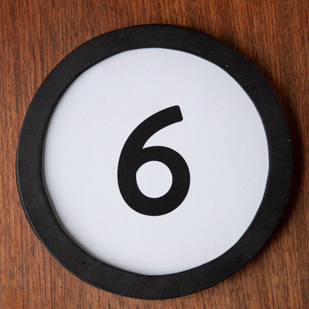 A circle shaped token fills the square image. The token has a 1 inch black circular frame around it. In the center is a black number on a white background. The token lies on a teak wood table. This digital image is part of the 1 to Infinity portrait photography series by Danny Goldfield.