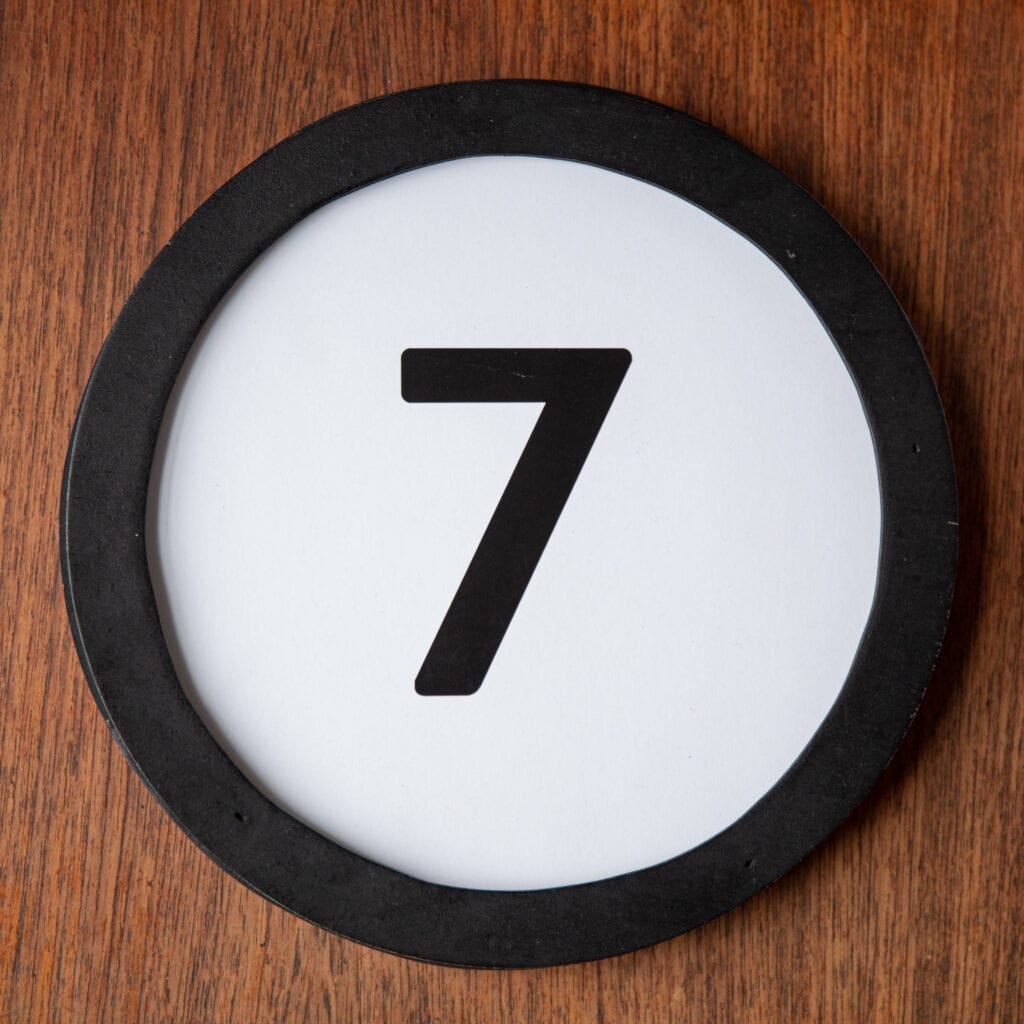 A circle shaped token fills the square image. The token has a 1 inch black circular frame around it. In the center is a black number on a white background. The token lies on a teak wood table. This digital image is part of the 1 to Infinity portrait photography series by Danny Goldfield.