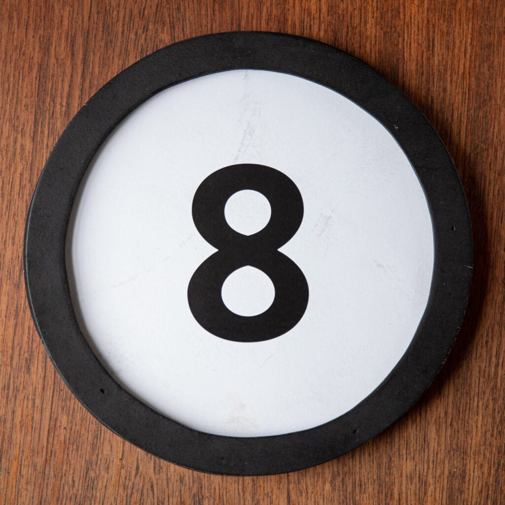 A circle shaped token fills the square image. The token has a 1 inch black circular frame around it. In the center is a black number on a white background. The token lies on a teak wood table. This digital image is part of the 1 to Infinity portrait photography series by Danny Goldfield.