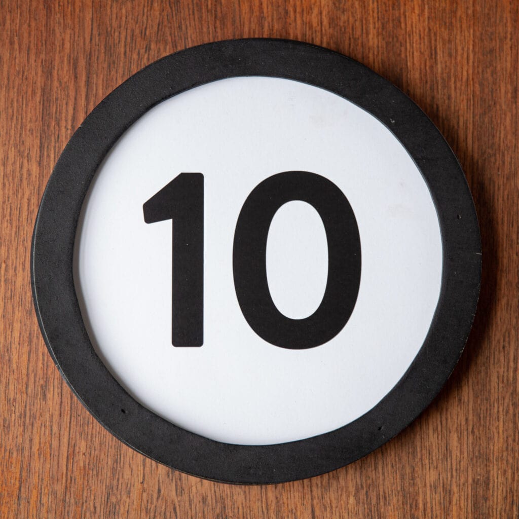 A circle shaped token fills the square image. The token has a 1 inch black circular frame around it. In the center is a black number on a white background. The token lies on a teak wood table. This digital image is part of the 1 to Infinity portrait photography series by Danny Goldfield.