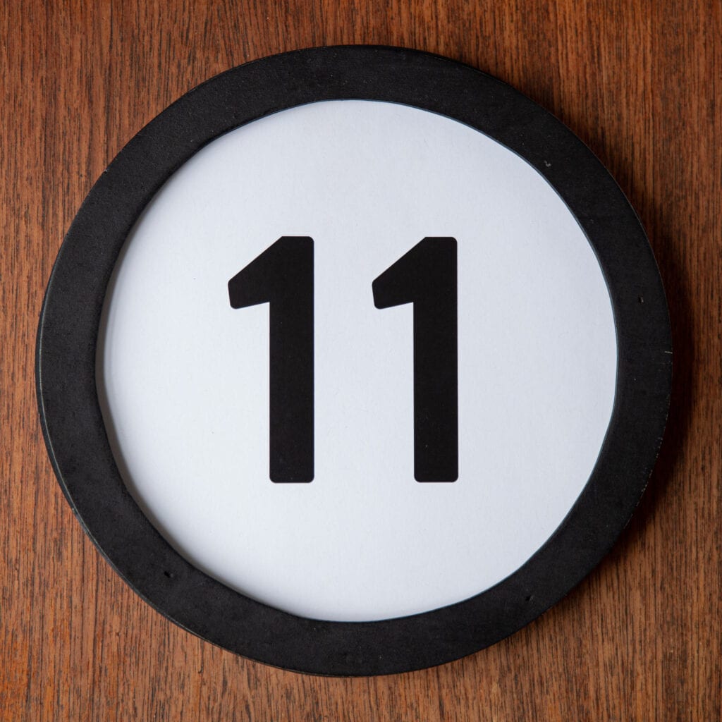 A circle shaped token fills the square image. The token has a 1 inch black circular frame around it. In the center is a black number on a white background. The token lies on a teak wood table. This digital image is part of the 1 to Infinity portrait photography series by Danny Goldfield.