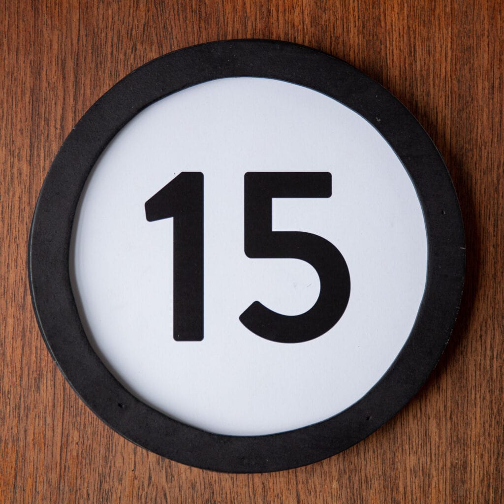 A circle shaped token fills the square image. The token has a 1 inch black circular frame around it. In the center is a black number on a white background. The token lies on a teak wood table. This digital image is part of the 1 to Infinity portrait photography series by Danny Goldfield.