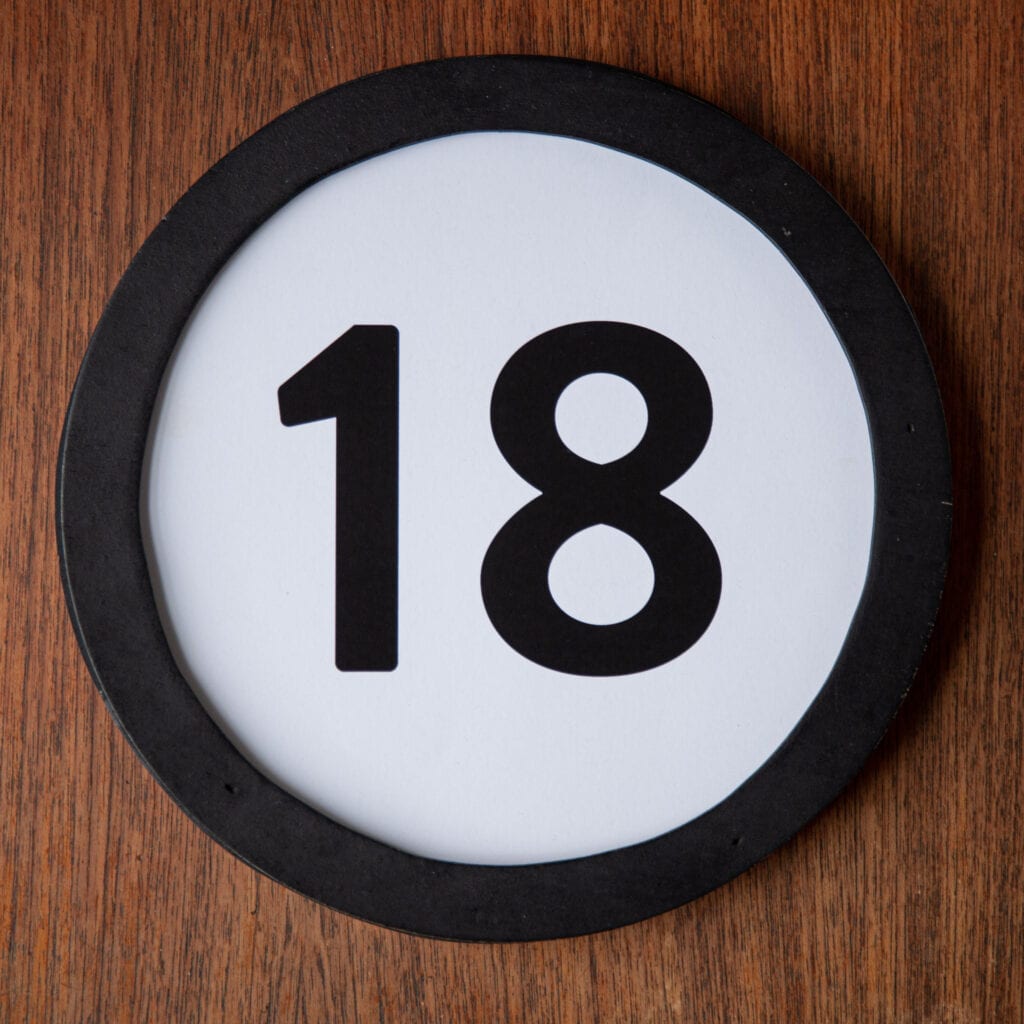 A circle shaped token fills the square image. The token has a 1 inch black circular frame around it. In the center is a black number on a white background. The token lies on a teak wood table. This digital image is part of the 1 to Infinity portrait photography series by Danny Goldfield.