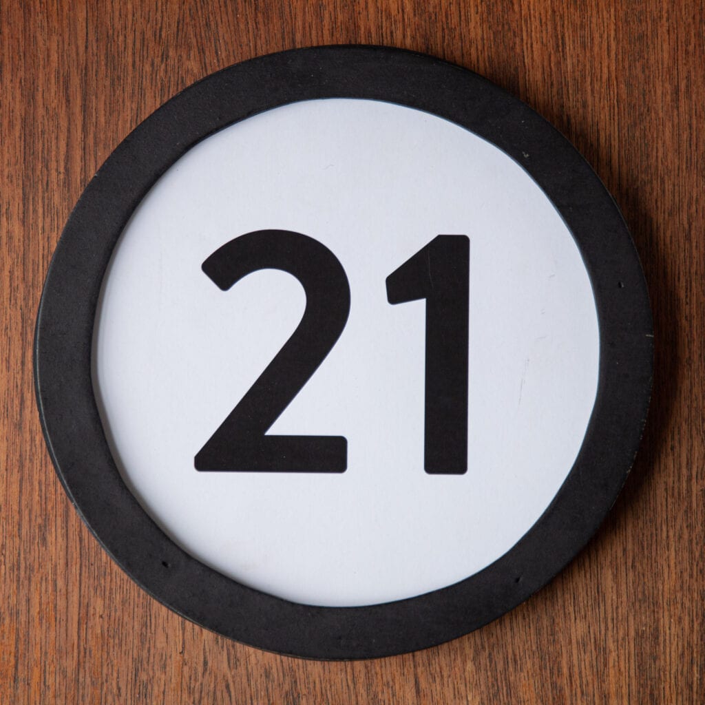 A circle shaped token fills the square image. The token has a 1 inch black circular frame around it. In the center is a black number on a white background. The token lies on a teak wood table. This digital image is part of the 1 to Infinity portrait photography series by Danny Goldfield.