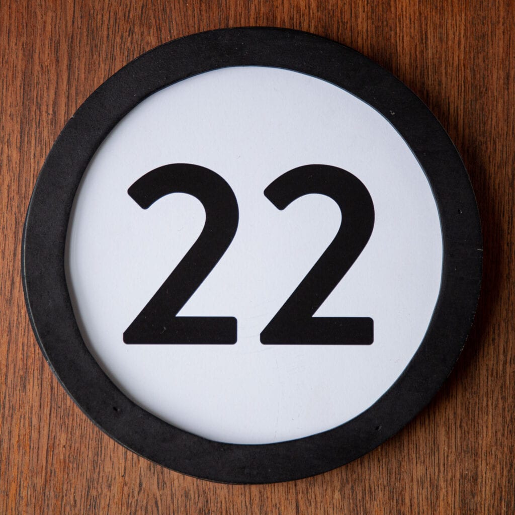 A circle shaped token fills the square image. The token has a 1 inch black circular frame around it. In the center is a black number on a white background. The token lies on a teak wood table. This digital image is part of the 1 to Infinity portrait photography series by Danny Goldfield.