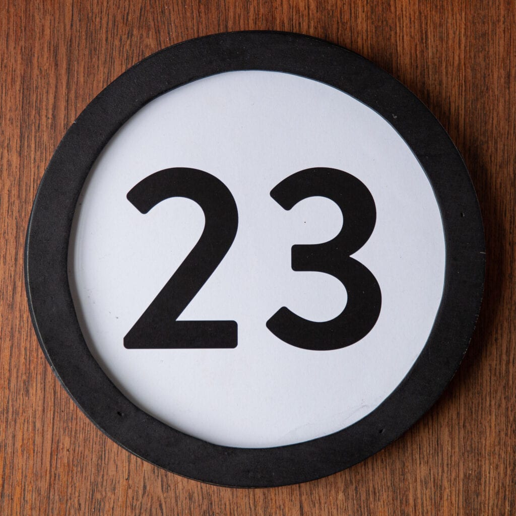 A circle shaped token fills the square image. The token has a 1 inch black circular frame around it. In the center is a black number on a white background. The token lies on a teak wood table. This digital image is part of the 1 to Infinity portrait photography series by Danny Goldfield.