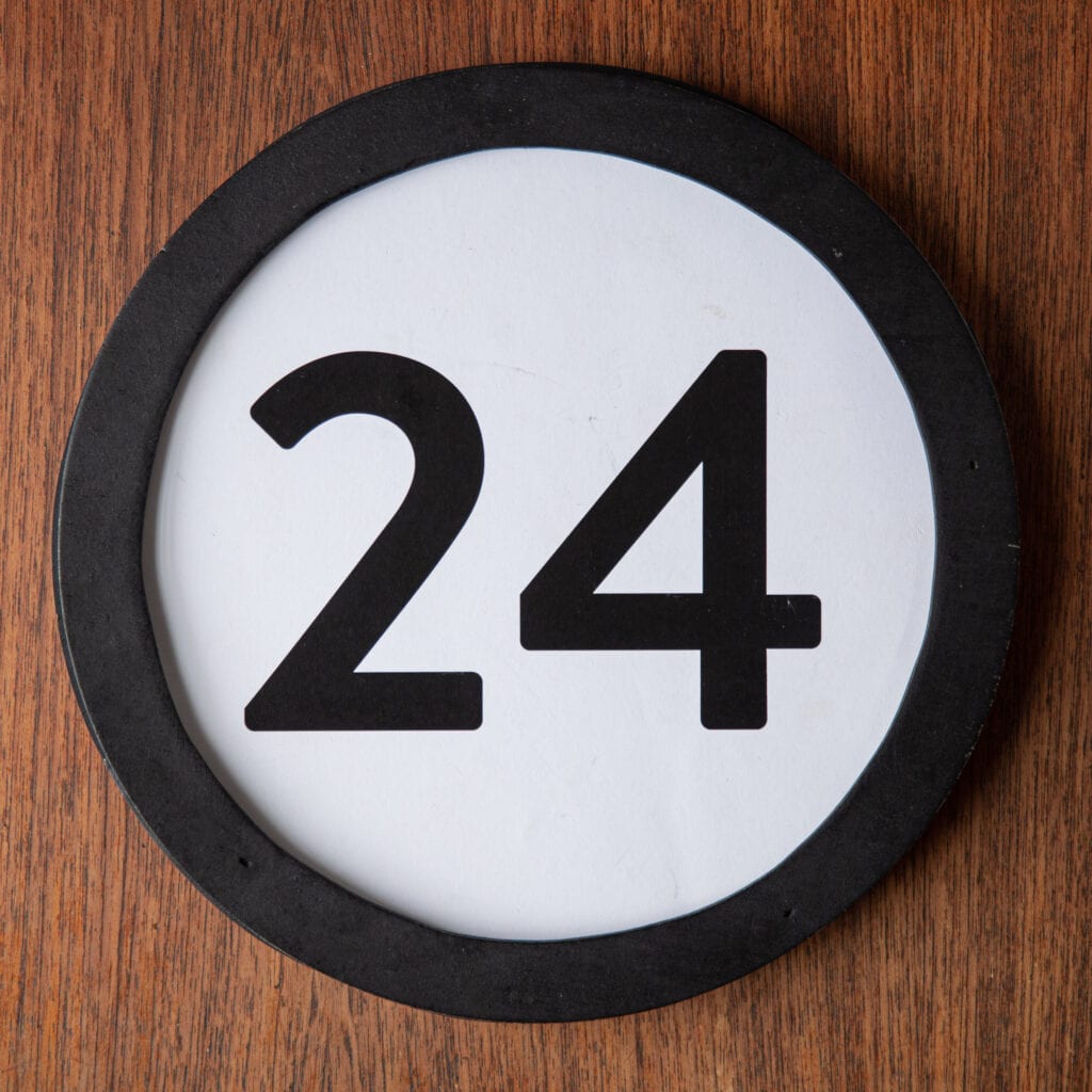 A circle shaped token fills the square image. The token has a 1 inch black circular frame around it. In the center is a black number on a white background. The token lies on a teak wood table. This digital image is part of the 1 to Infinity portrait photography series by Danny Goldfield.