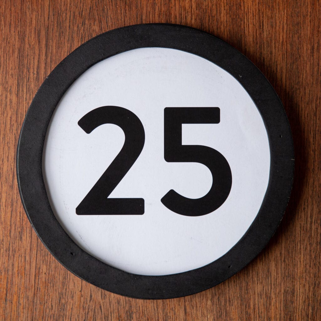 A circle shaped token fills the square image. The token has a 1 inch black circular frame around it. In the center is a black number on a white background. The token lies on a teak wood table. This digital image is part of the 1 to Infinity portrait photography series by Danny Goldfield.