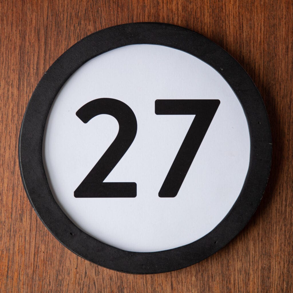 A circle shaped token fills the square image. The token has a 1 inch black circular frame around it. In the center is a black number on a white background. The token lies on a teak wood table. This digital image is part of the 1 to Infinity portrait photography series by Danny Goldfield.