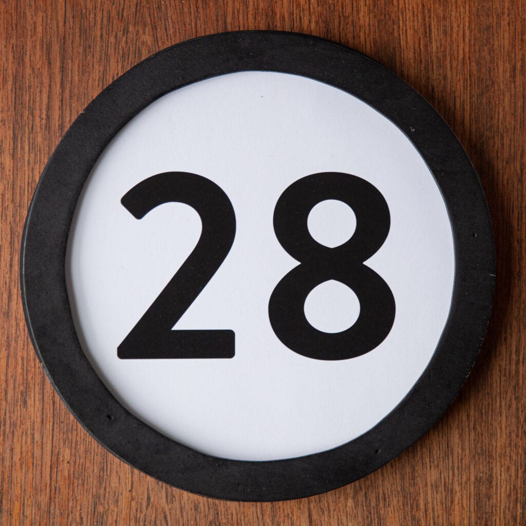 A circle shaped token fills the square image. The token has a 1 inch black circular frame around it. In the center is a black number on a white background. The token lies on a teak wood table. This digital image is part of the 1 to Infinity portrait photography series by Danny Goldfield.