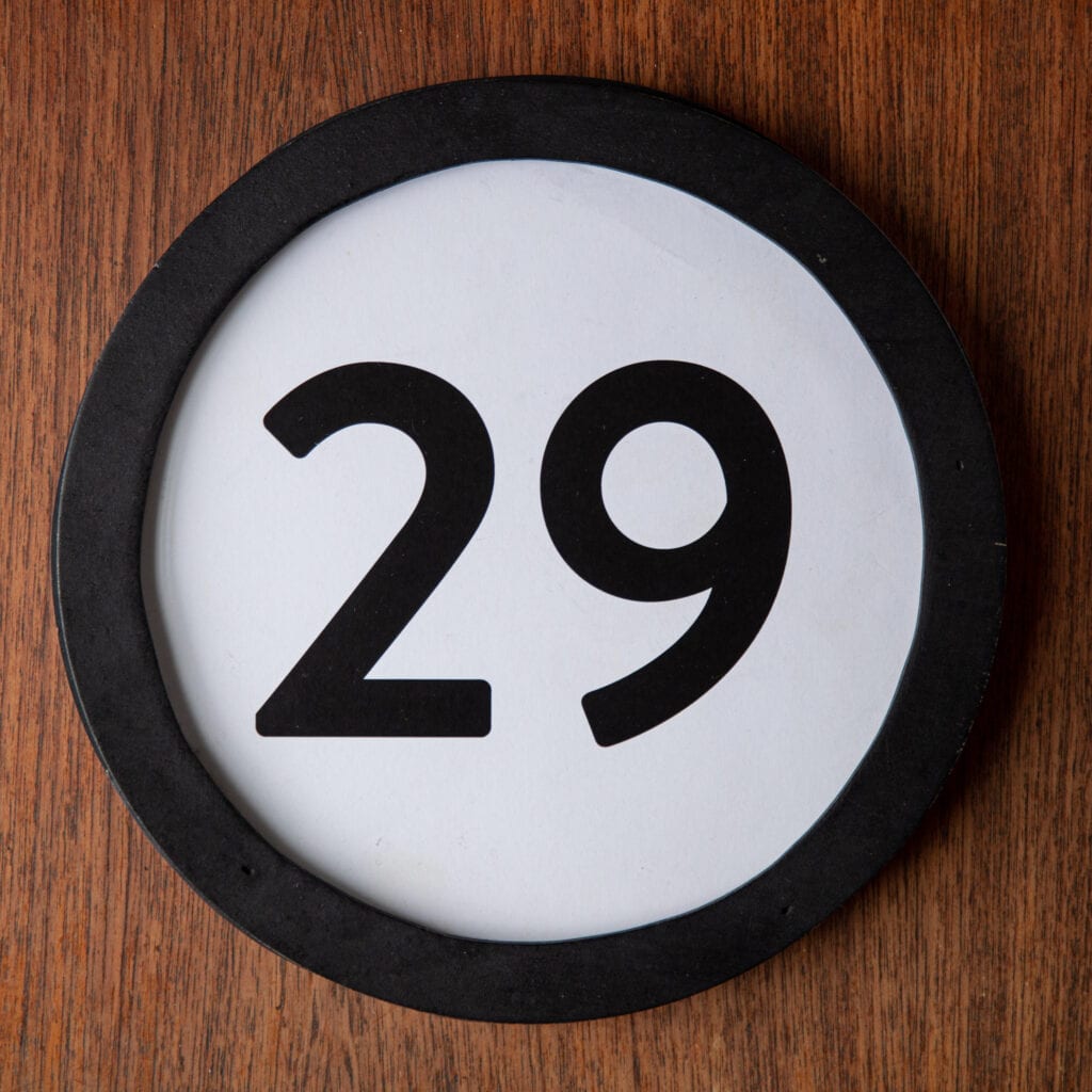 A circle shaped token fills the square image. The token has a 1 inch black circular frame around it. In the center is a black number on a white background. The token lies on a teak wood table. This digital image is part of the 1 to Infinity portrait photography series by Danny Goldfield.