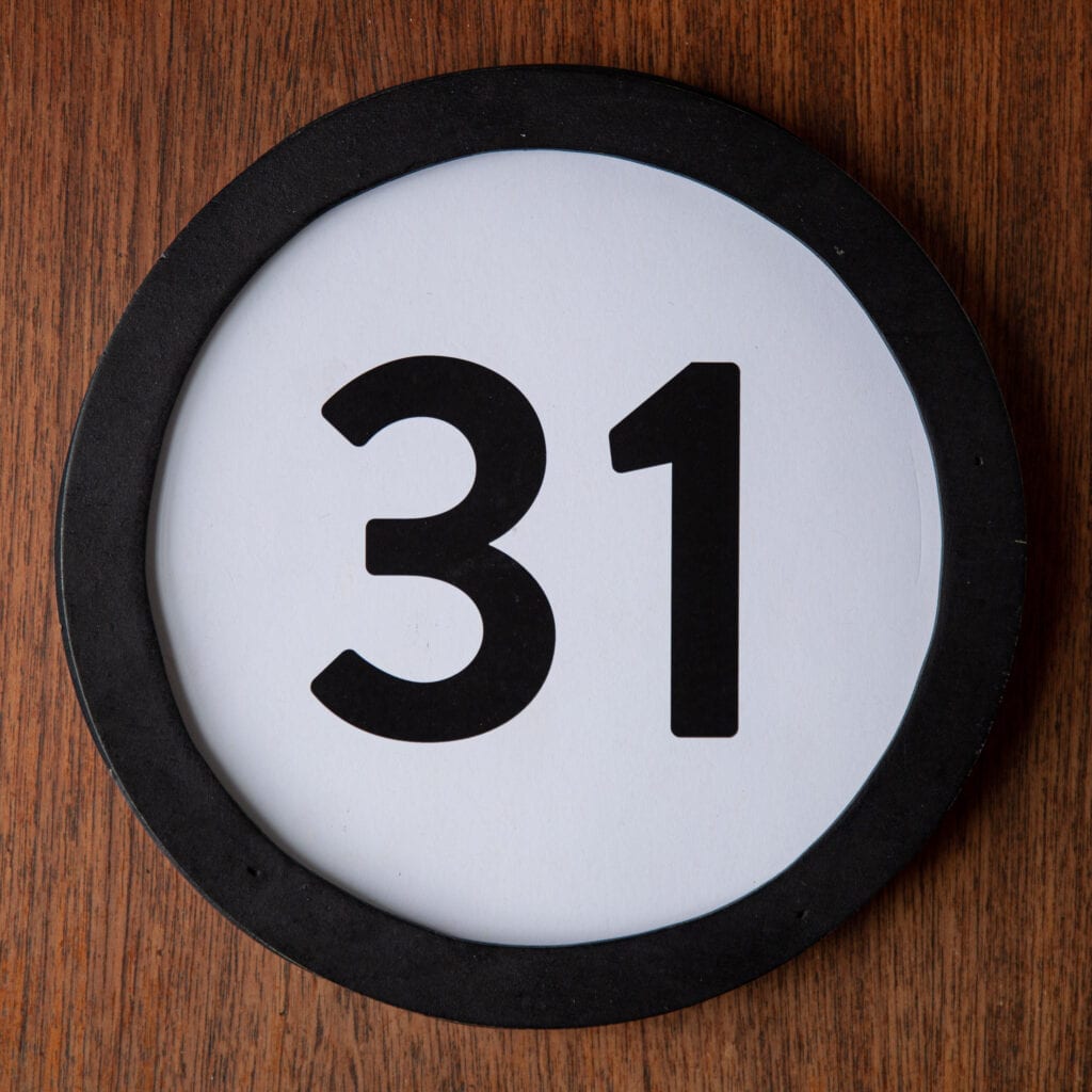A circle shaped token fills the square image. The token has a 1 inch black circular frame around it. In the center is a black number on a white background. The token lies on a teak wood table. This digital image is part of the 1 to Infinity portrait photography series by Danny Goldfield.