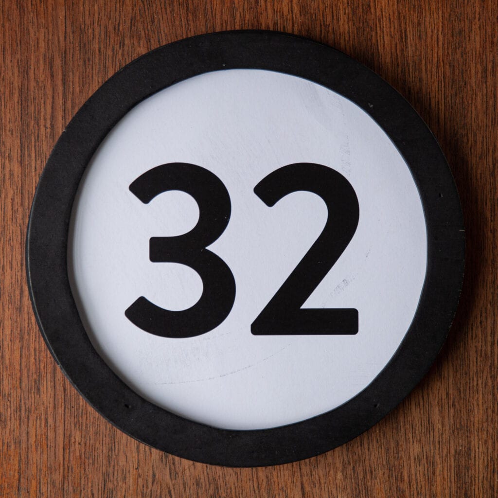 A circle shaped token fills the square image. The token has a 1 inch black circular frame around it. In the center is a black number on a white background. The token lies on a teak wood table. This digital image is part of the 1 to Infinity portrait photography series by Danny Goldfield.