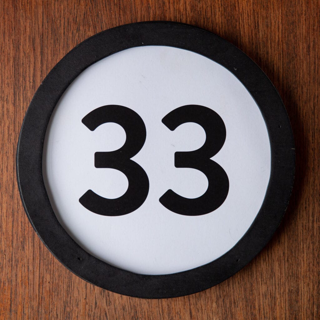 A circle shaped token fills the square image. The token has a 1 inch black circular frame around it. In the center is a black number on a white background. The token lies on a teak wood table. This digital image is part of the 1 to Infinity portrait photography series by Danny Goldfield.
