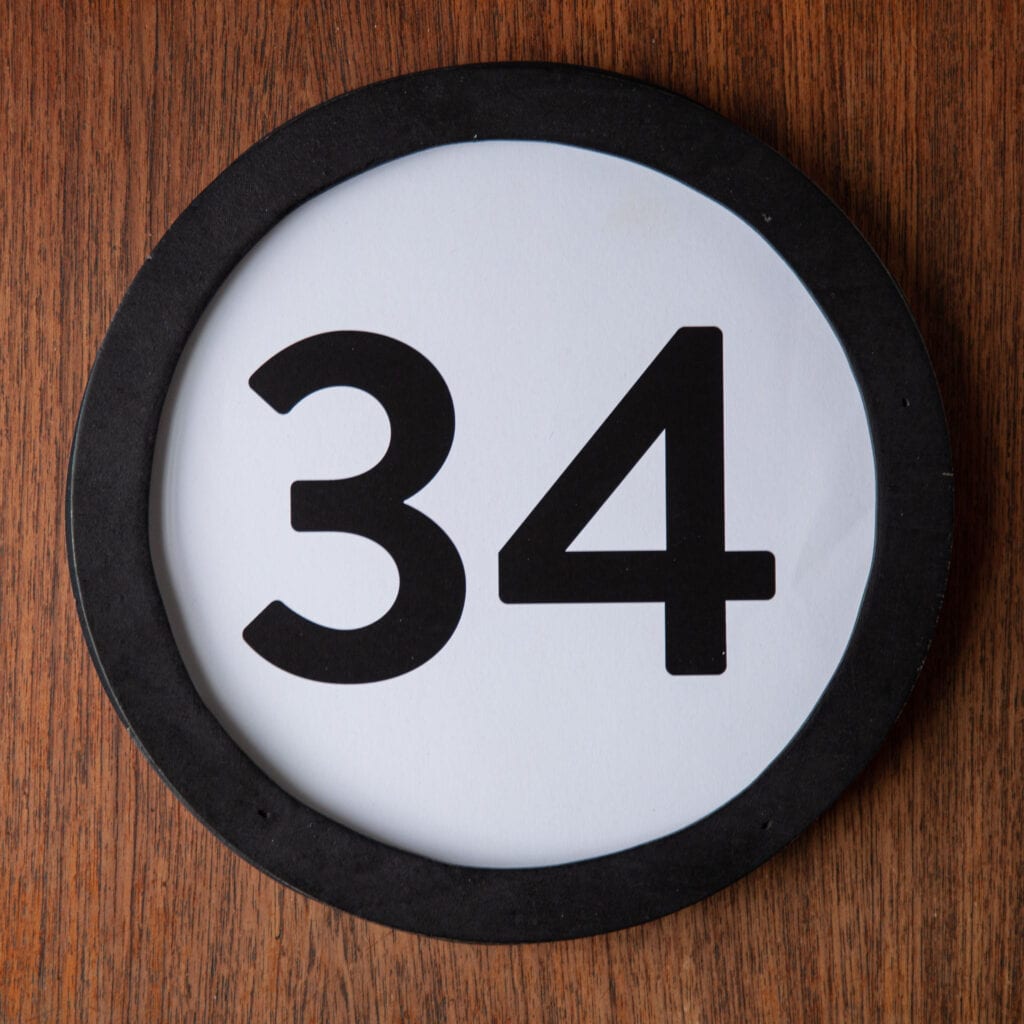 A circle shaped token fills the square image. The token has a 1 inch black circular frame around it. In the center is a black number on a white background. The token lies on a teak wood table. This digital image is part of the 1 to Infinity portrait photography series by Danny Goldfield.