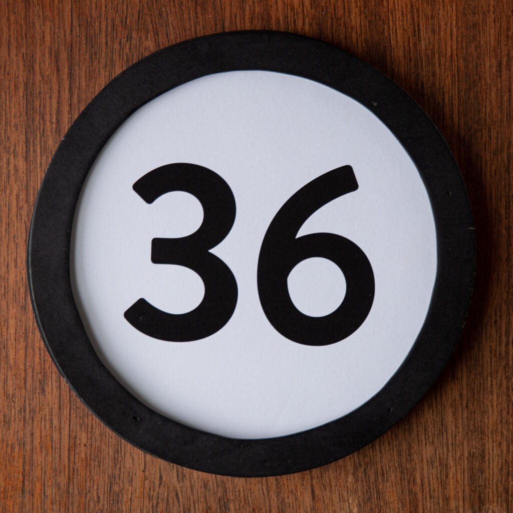 A circle shaped token fills the square image. The token has a 1 inch black circular frame around it. In the center is a black number on a white background. The token lies on a teak wood table. This digital image is part of the 1 to Infinity portrait photography series by Danny Goldfield.