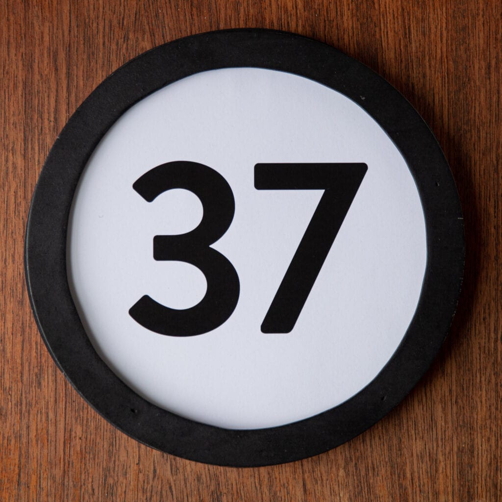 A circle shaped token fills the square image. The token has a 1 inch black circular frame around it. In the center is a black number on a white background. The token lies on a teak wood table. This digital image is part of the 1 to Infinity portrait photography series by Danny Goldfield.