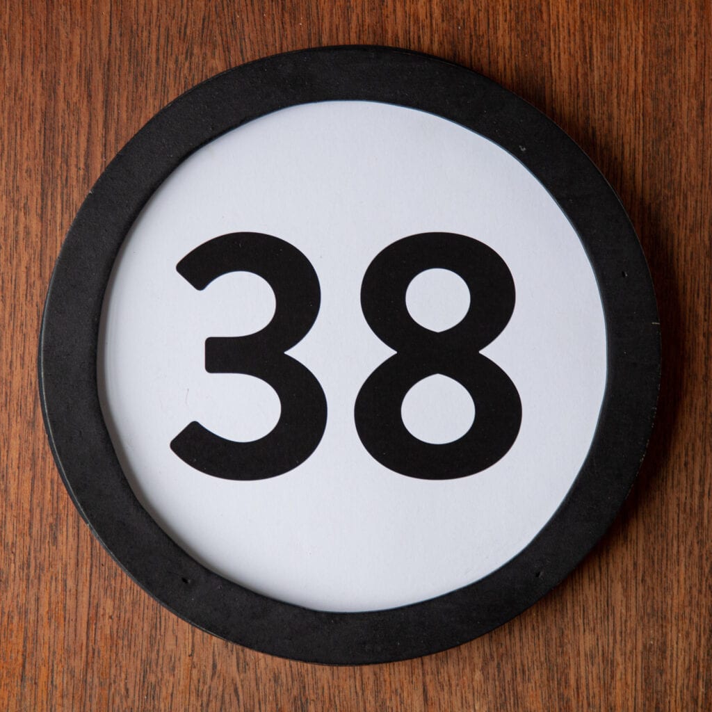 A circle shaped token fills the square image. The token has a 1 inch black circular frame around it. In the center is a black number on a white background. The token lies on a teak wood table. This digital image is part of the 1 to Infinity portrait photography series by Danny Goldfield.