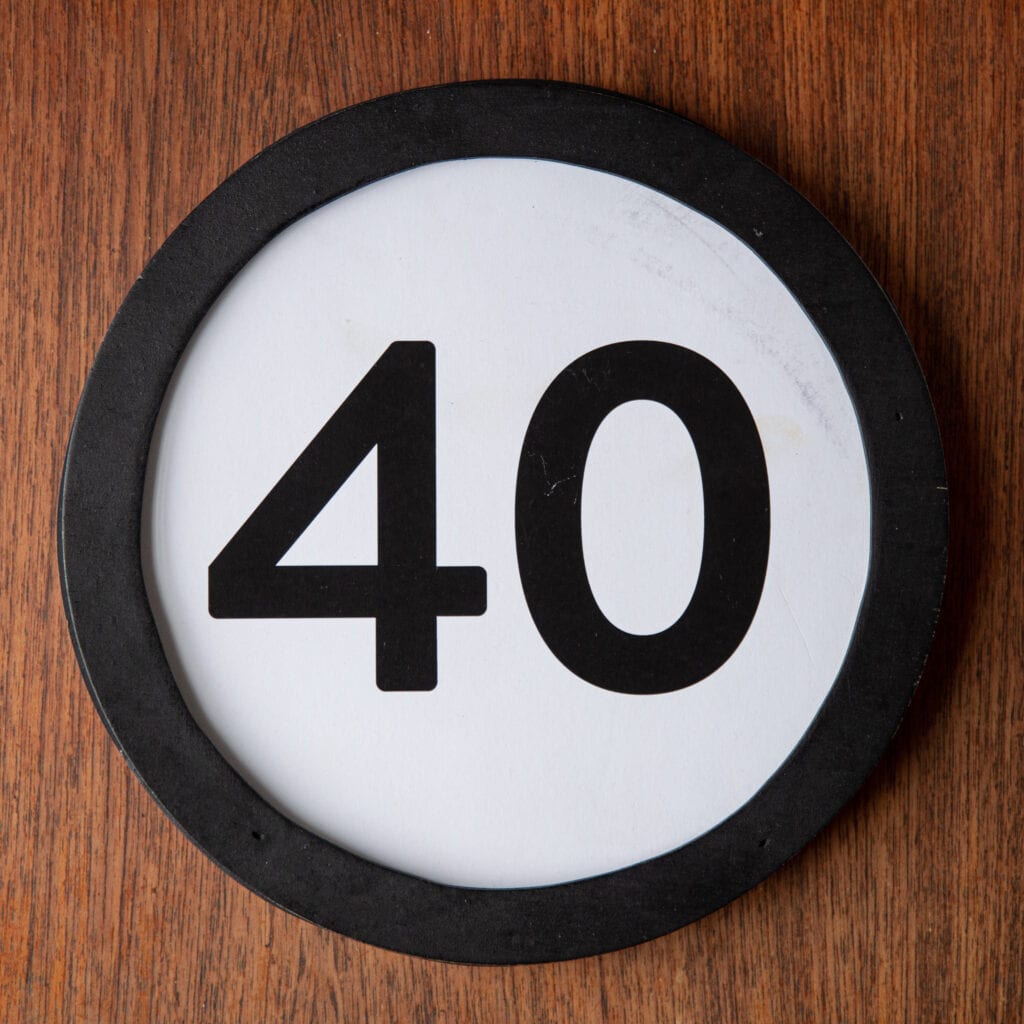 A circle shaped token fills the square image. The token has a 1 inch black circular frame around it. In the center is a black number on a white background. The token lies on a teak wood table. This digital image is part of the 1 to Infinity portrait photography series by Danny Goldfield.