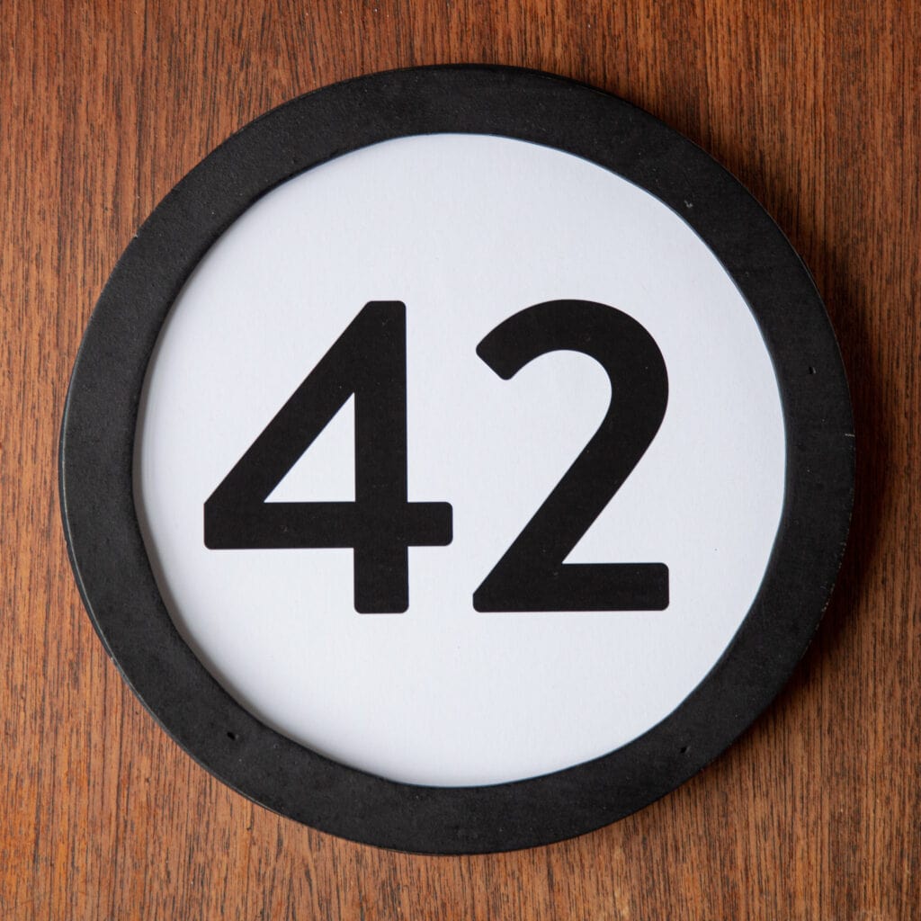 A circle shaped token fills the square image. The token has a 1 inch black circular frame around it. In the center is a black number on a white background. The token lies on a teak wood table. This digital image is part of the 1 to Infinity portrait photography series by Danny Goldfield.