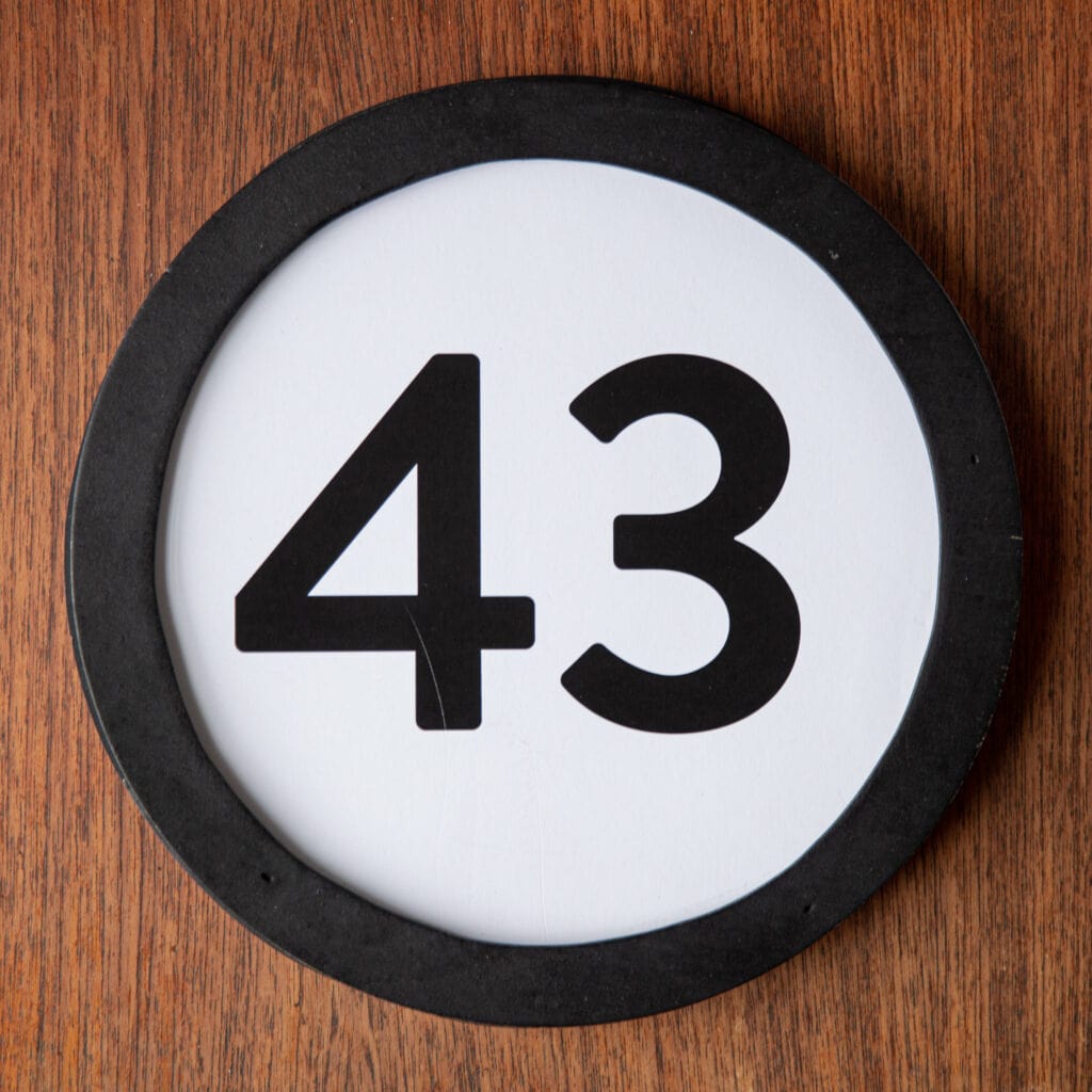 A circle shaped token fills the square image. The token has a 1 inch black circular frame around it. In the center is a black number on a white background. The token lies on a teak wood table. This digital image is part of the 1 to Infinity portrait photography series by Danny Goldfield.