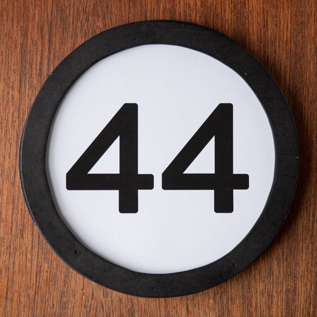 A circle shaped token fills the square image. The token has a 1 inch black circular frame around it. In the center is a black number on a white background. The token lies on a teak wood table. This digital image is part of the 1 to Infinity portrait photography series by Danny Goldfield.