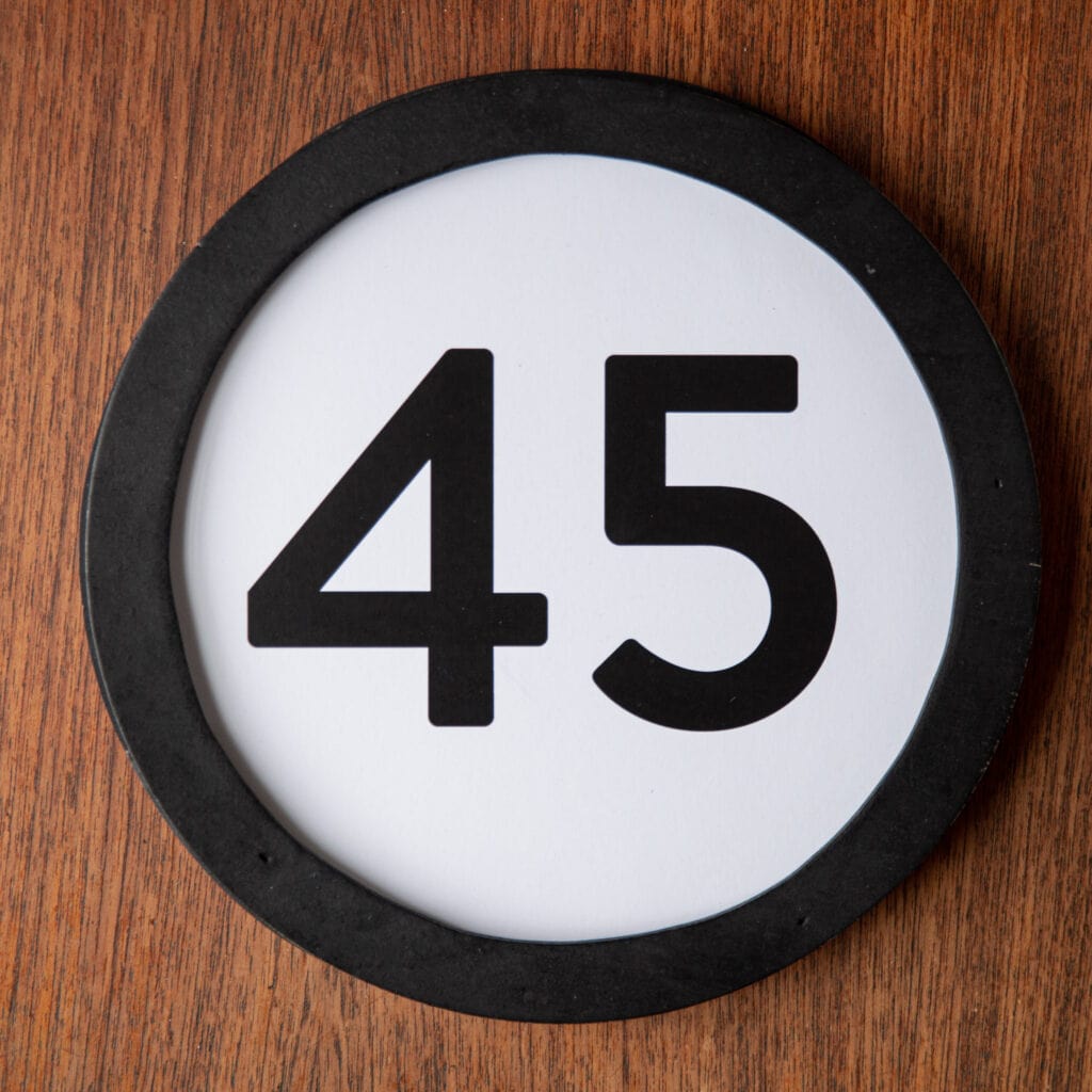 A circle shaped token fills the square image. The token has a 1 inch black circular frame around it. In the center is a black number on a white background. The token lies on a teak wood table. This digital image is part of the 1 to Infinity portrait photography series by Danny Goldfield.