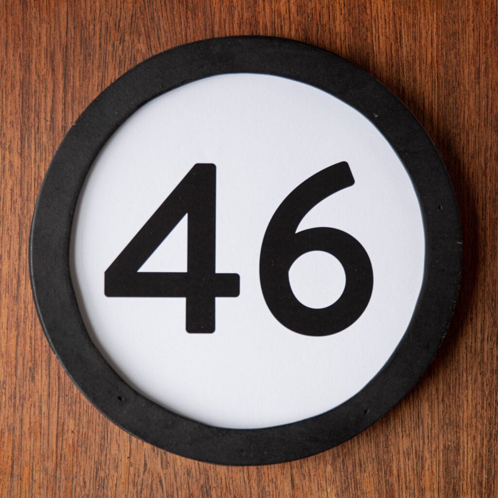A circle shaped token fills the square image. The token has a 1 inch black circular frame around it. In the center is a black number on a white background. The token lies on a teak wood table. This digital image is part of the 1 to Infinity portrait photography series by Danny Goldfield.