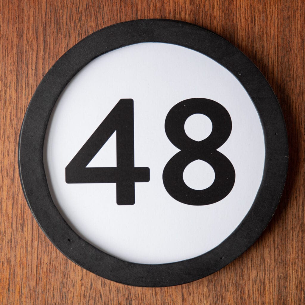 A circle shaped token fills the square image. The token has a 1 inch black circular frame around it. In the center is a black number on a white background. The token lies on a teak wood table. This digital image is part of the 1 to Infinity portrait photography series by Danny Goldfield.