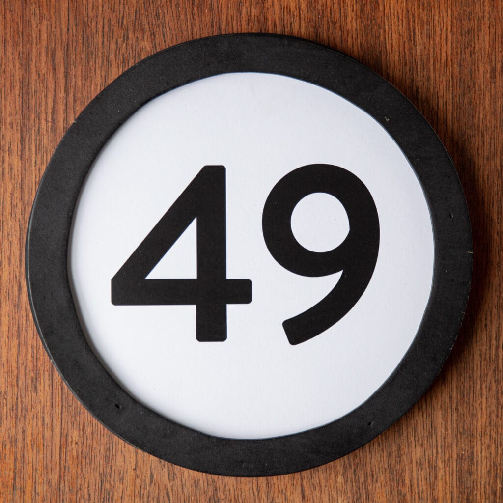 A circle shaped token fills the square image. The token has a 1 inch black circular frame around it. In the center is a black number on a white background. The token lies on a teak wood table. This digital image is part of the 1 to Infinity portrait photography series by Danny Goldfield.