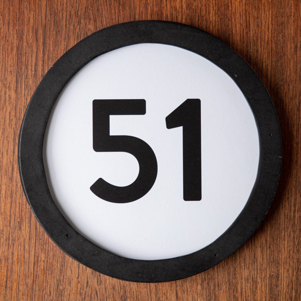 A circle shaped token fills the square image. The token has a 1 inch black circular frame around it. In the center is a black number on a white background. The token lies on a teak wood table. This digital image is part of the 1 to Infinity portrait photography series by Danny Goldfield.