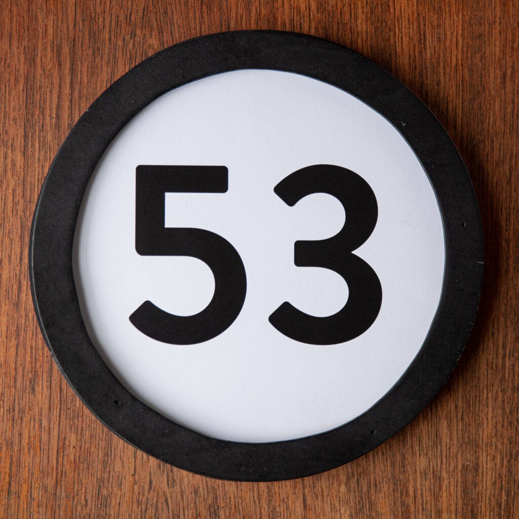 A circle shaped token fills the square image. The token has a 1 inch black circular frame around it. In the center is a black number on a white background. The token lies on a teak wood table. This digital image is part of the 1 to Infinity portrait photography series by Danny Goldfield.