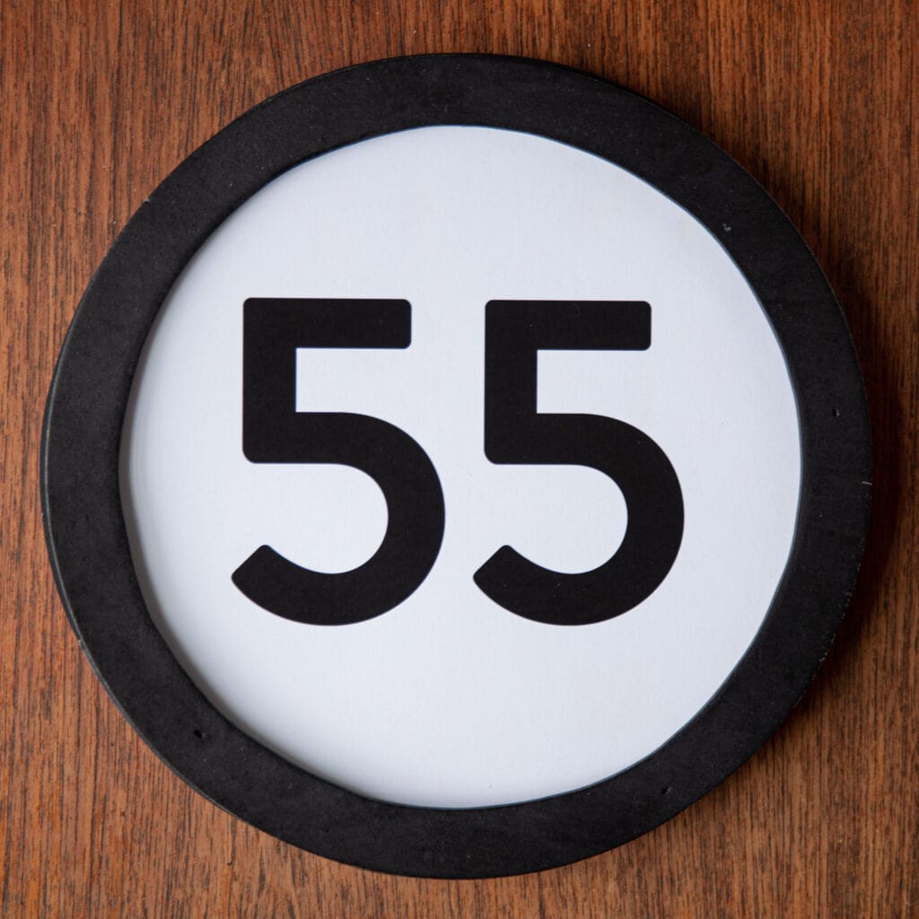A circle shaped token fills the square image. The token has a 1 inch black circular frame around it. In the center is a black number on a white background. The token lies on a teak wood table. This digital image is part of the 1 to Infinity portrait photography series by Danny Goldfield.